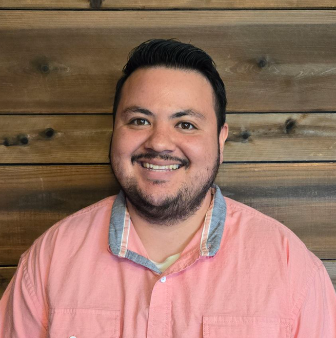 Smiling man in a pink shirt standing against a wooden plank wall.