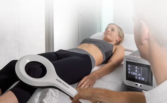 weißes medizinisches Gerät mit Touchscreen-Display und zwei Handsonden auf einem Ständer, neben einem Untersuchungstisch.