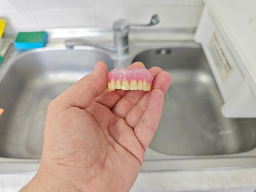 A Person Is Holding a Denture in Their Hand in Front of A Sink — Cosmetic Denture Services–Jacob Maxwell in Mackay, QLD