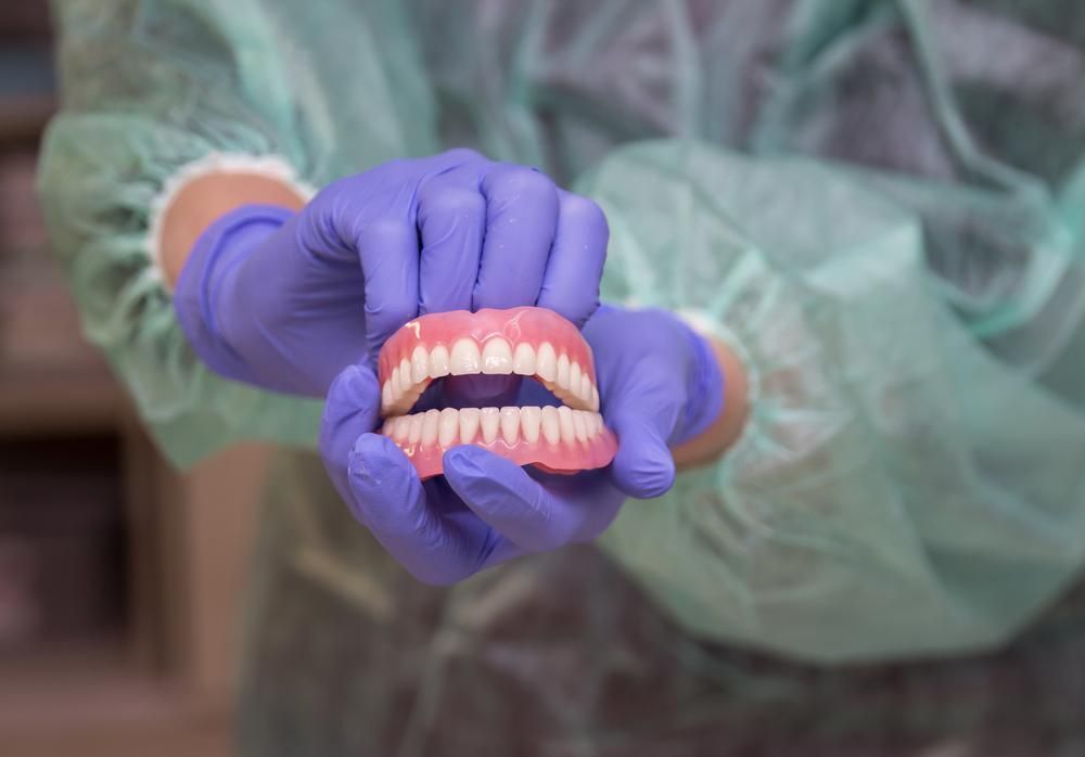 A Person Wearing Purple Gloves Is Holding a Denture in Their Hands — Cosmetic Denture Services–Jacob Maxwell in Mackay, QLD