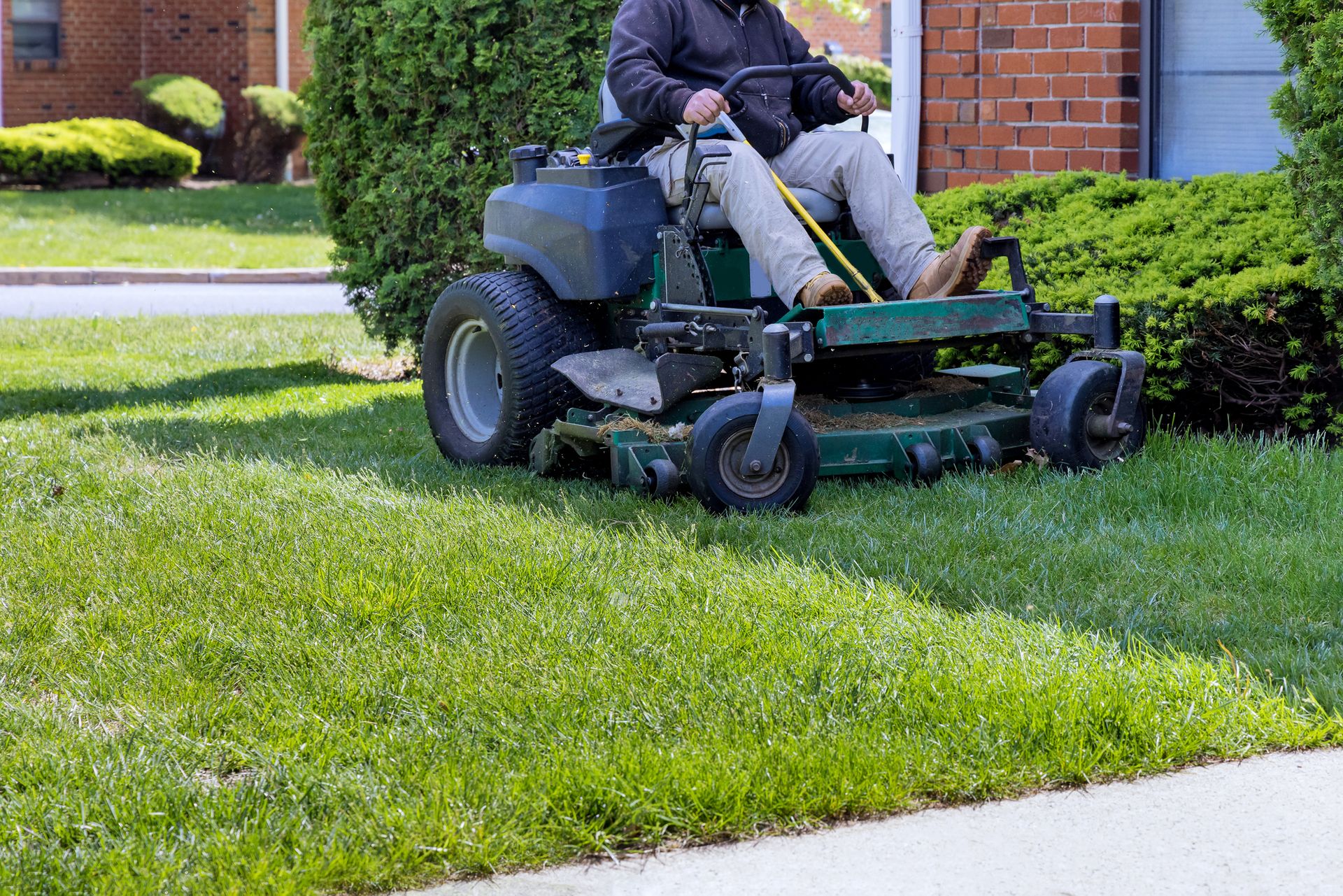 Person riding a green riding lawn mower on a grassy lawn beside a brick house