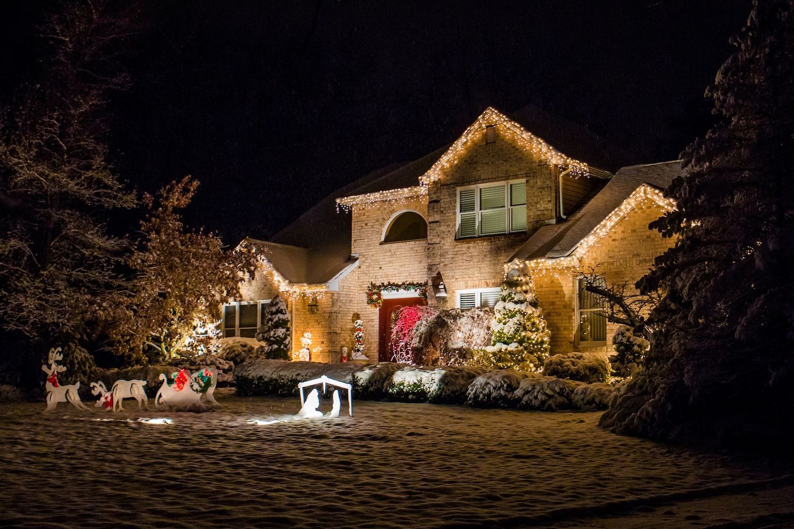 Snowy house glowing with holiday lights at night, with a red front door and festive decorations.