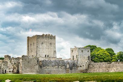 Stone castle ruins with a central tower under a cloudy sky, surrounded by grassy grounds.
