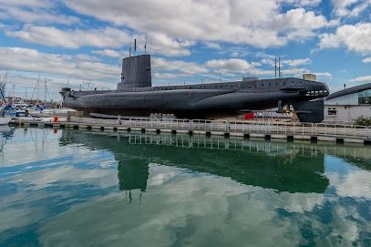 Submarine docked at a harbor, reflected in calm water under a cloudy sky.