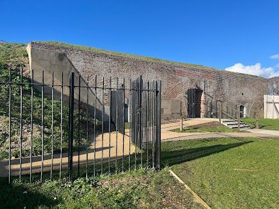 Stone fort wall with black metal fence, grassy foreground, and blue sky on a sunny day
