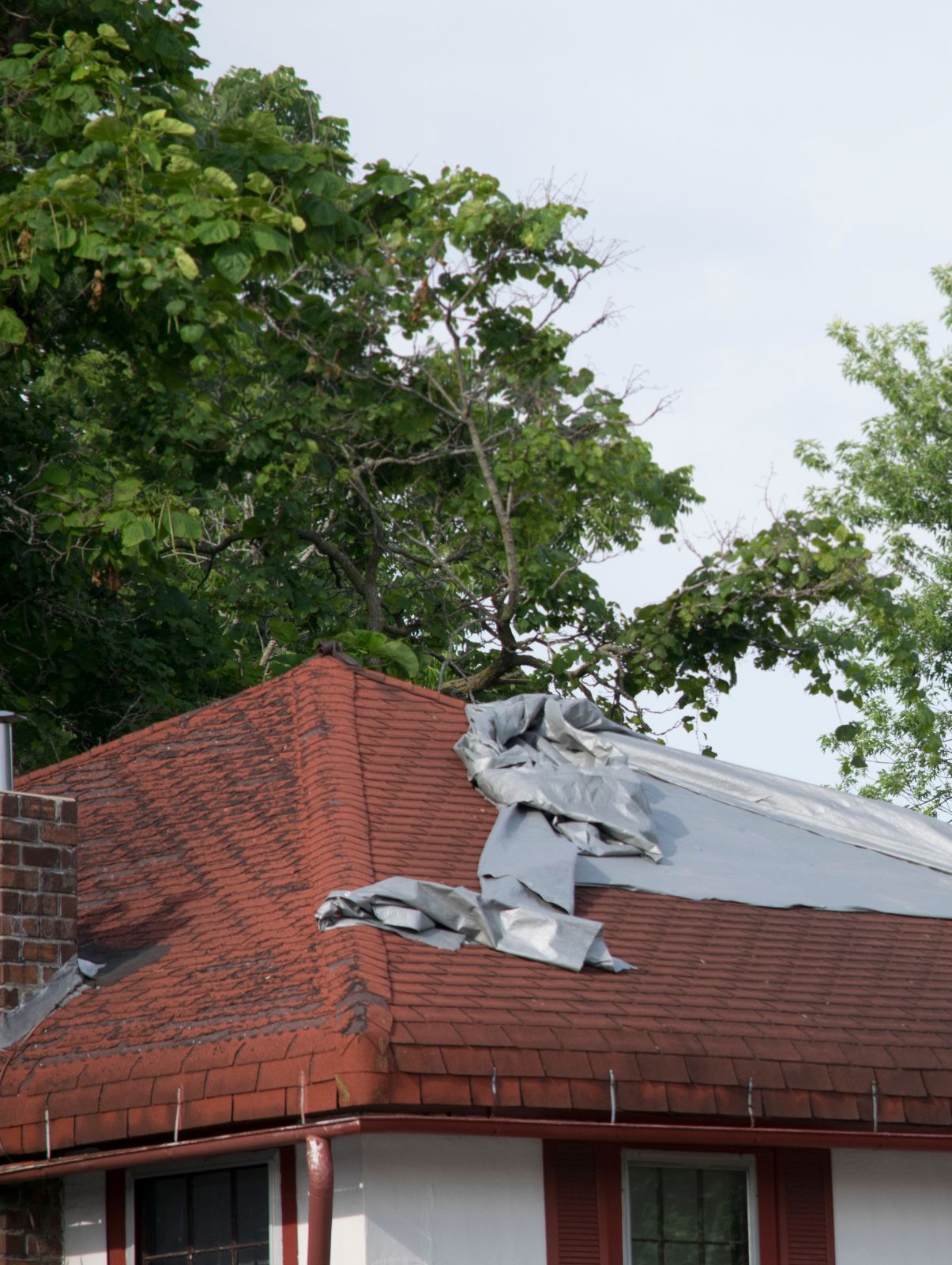 Damaged red roof with torn gray tarp, beside brick house and green trees