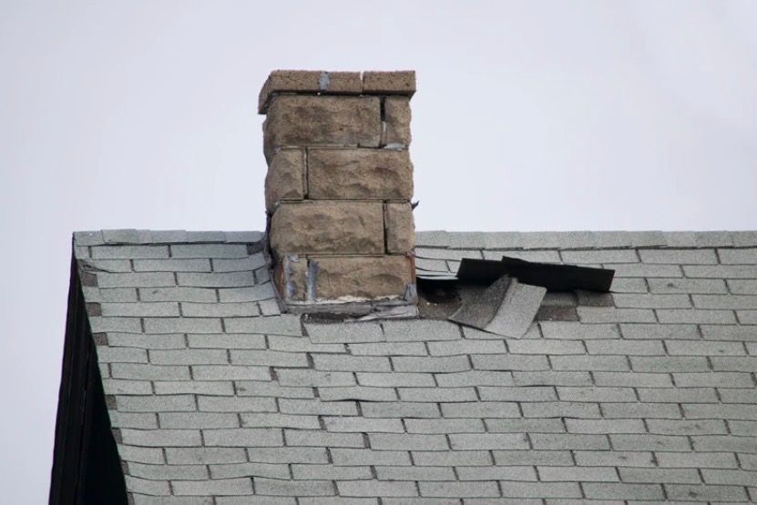 Damaged shingled roof with a stone chimney and missing shingles near the base
