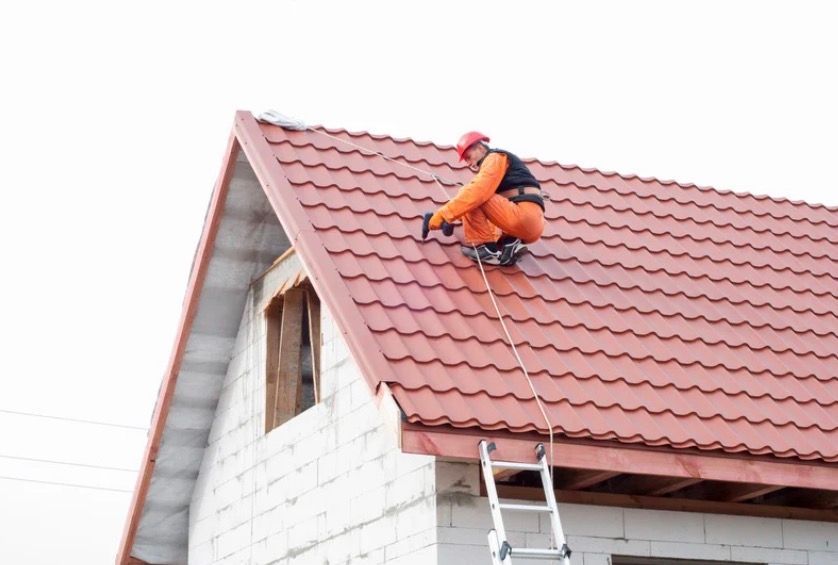 Worker in orange gear repairing a red shingled roof with a ladder below
