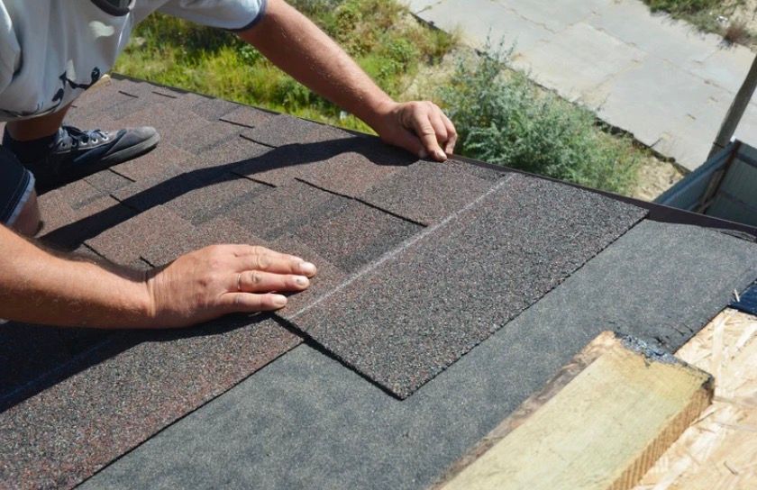 Person installing dark brown roof shingles on a sloped rooftop