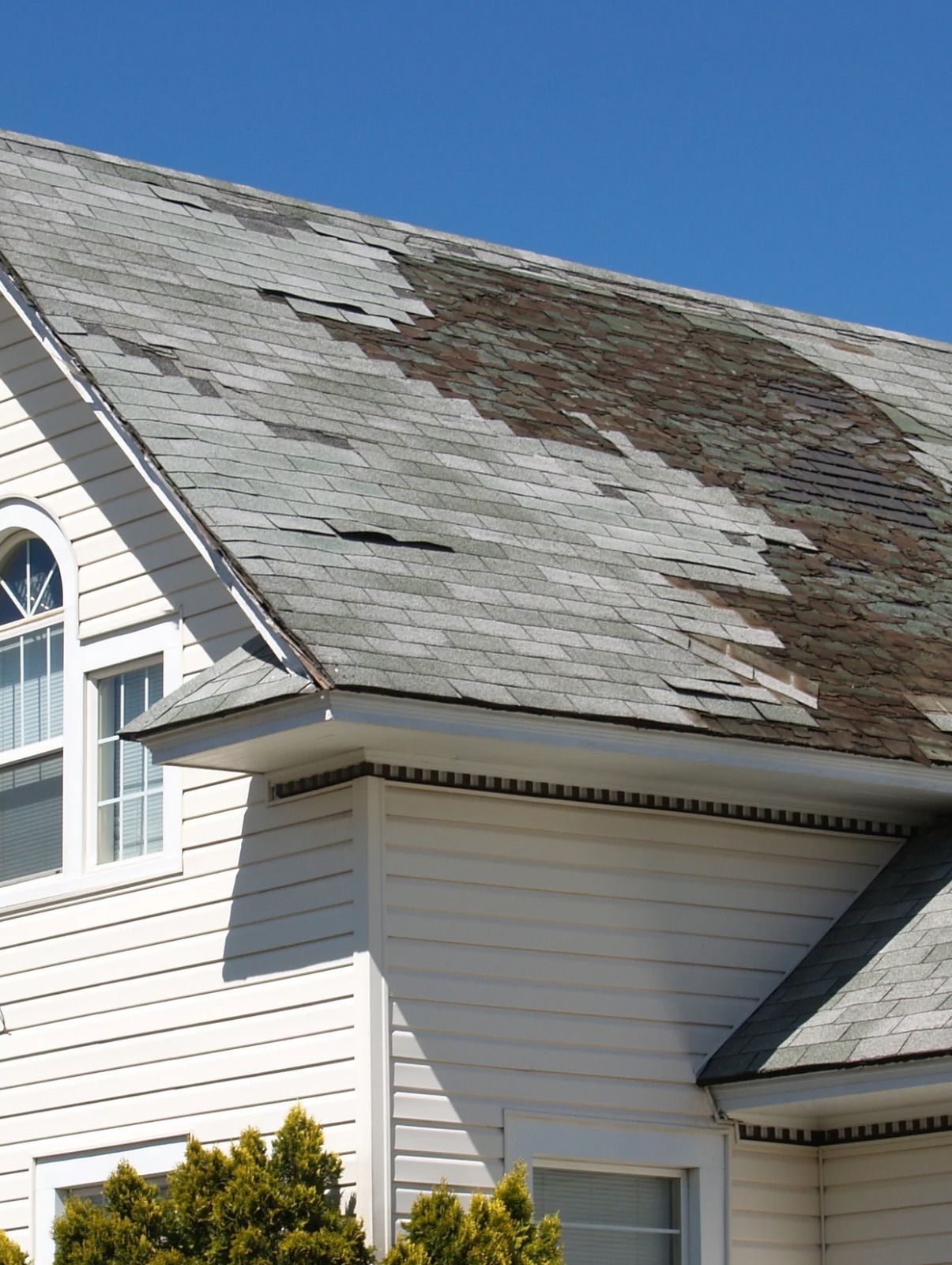 Damaged house roof with peeling shingles and large bare patch under a blue sky