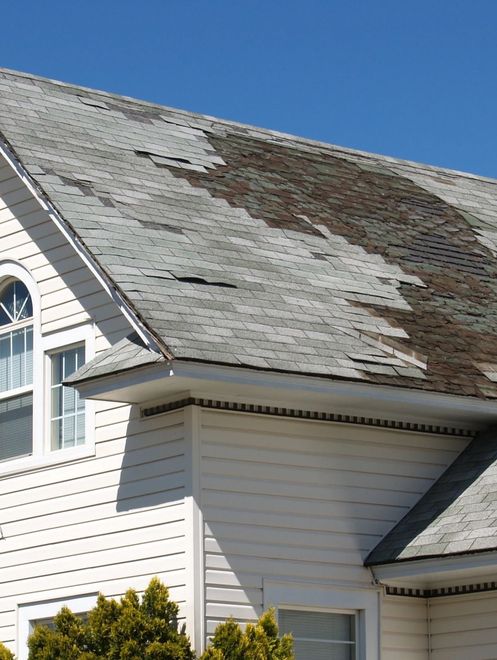 Damaged house roof with peeling shingles and large bare patch under a blue sky