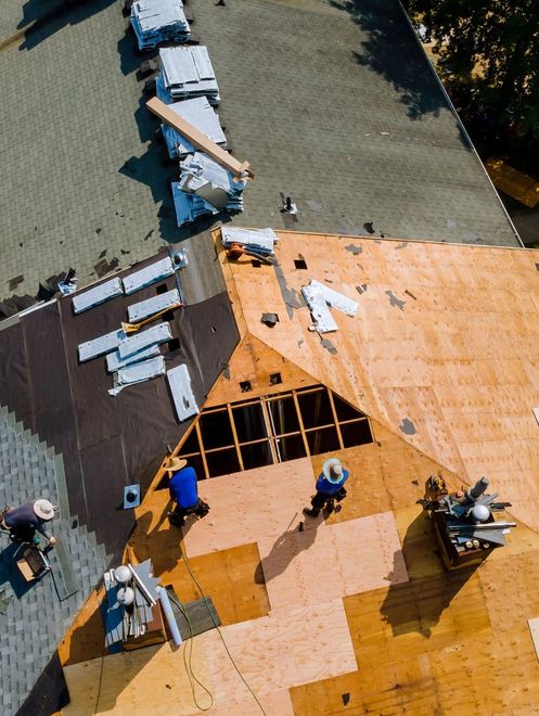 Roofers working on an orange roof, replacing damaged shingles around a large hole.