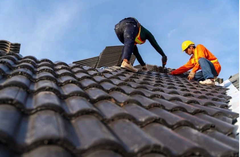 Two roof workers in safety gear repair gray tiled roof under a blue sky