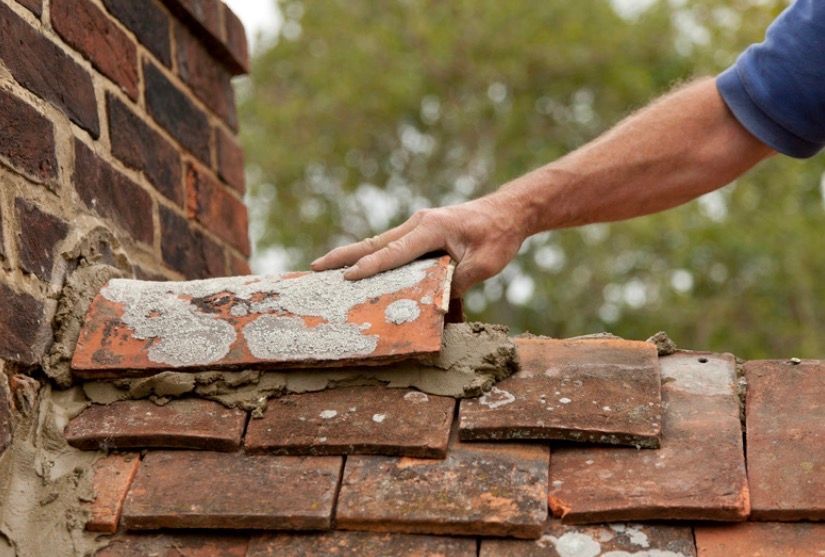 Hand placing a brick onto a partially rebuilt brick wall near a chimney