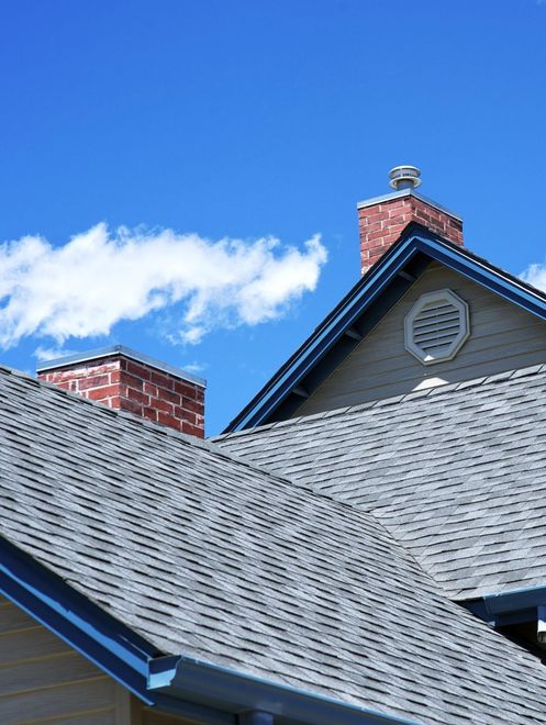 Residential roof with gray shingles and red brick chimneys under a blue sky with clouds