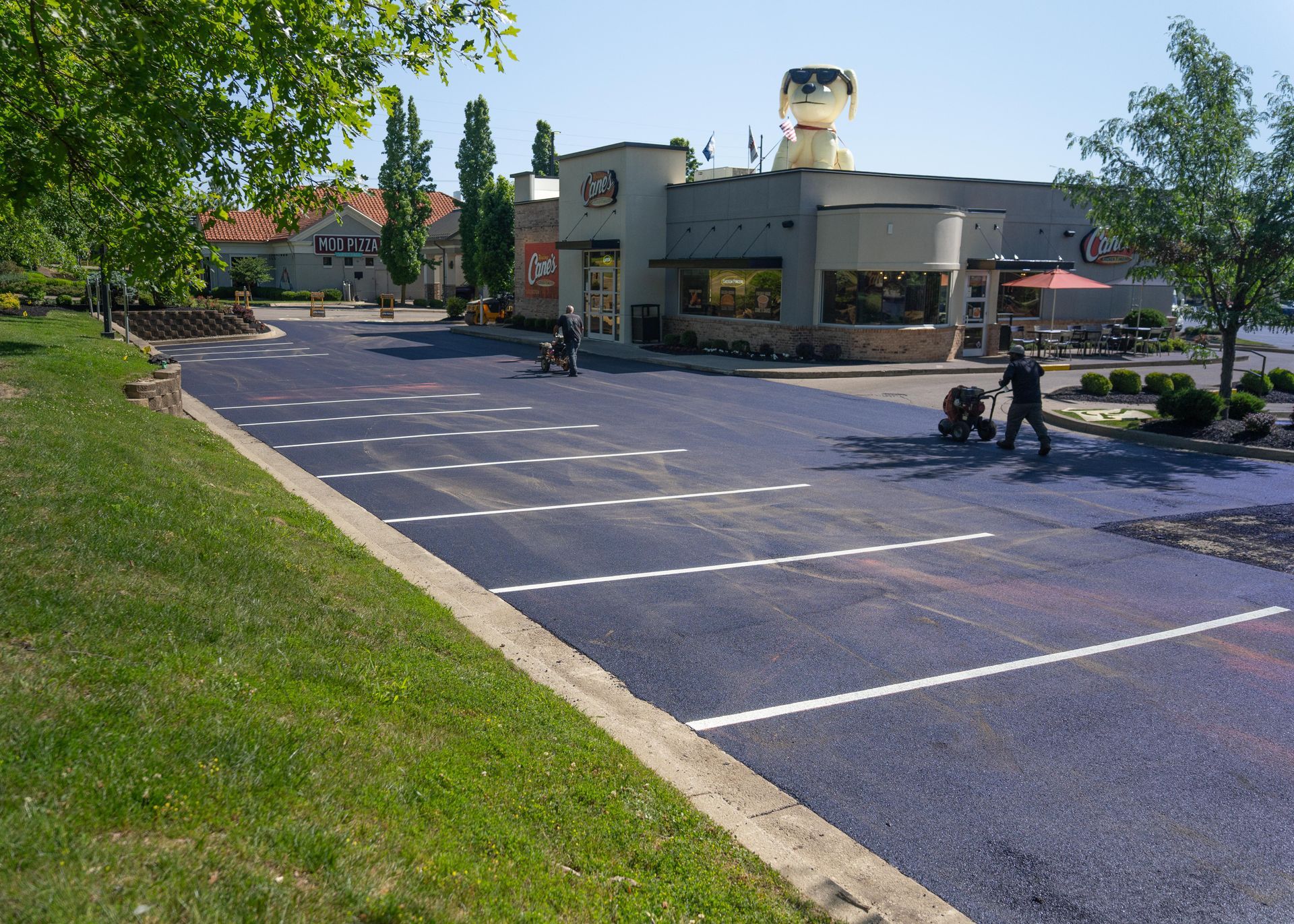 Parking lot beside a small building with a tree-lined lawn and two people walking near the entrance