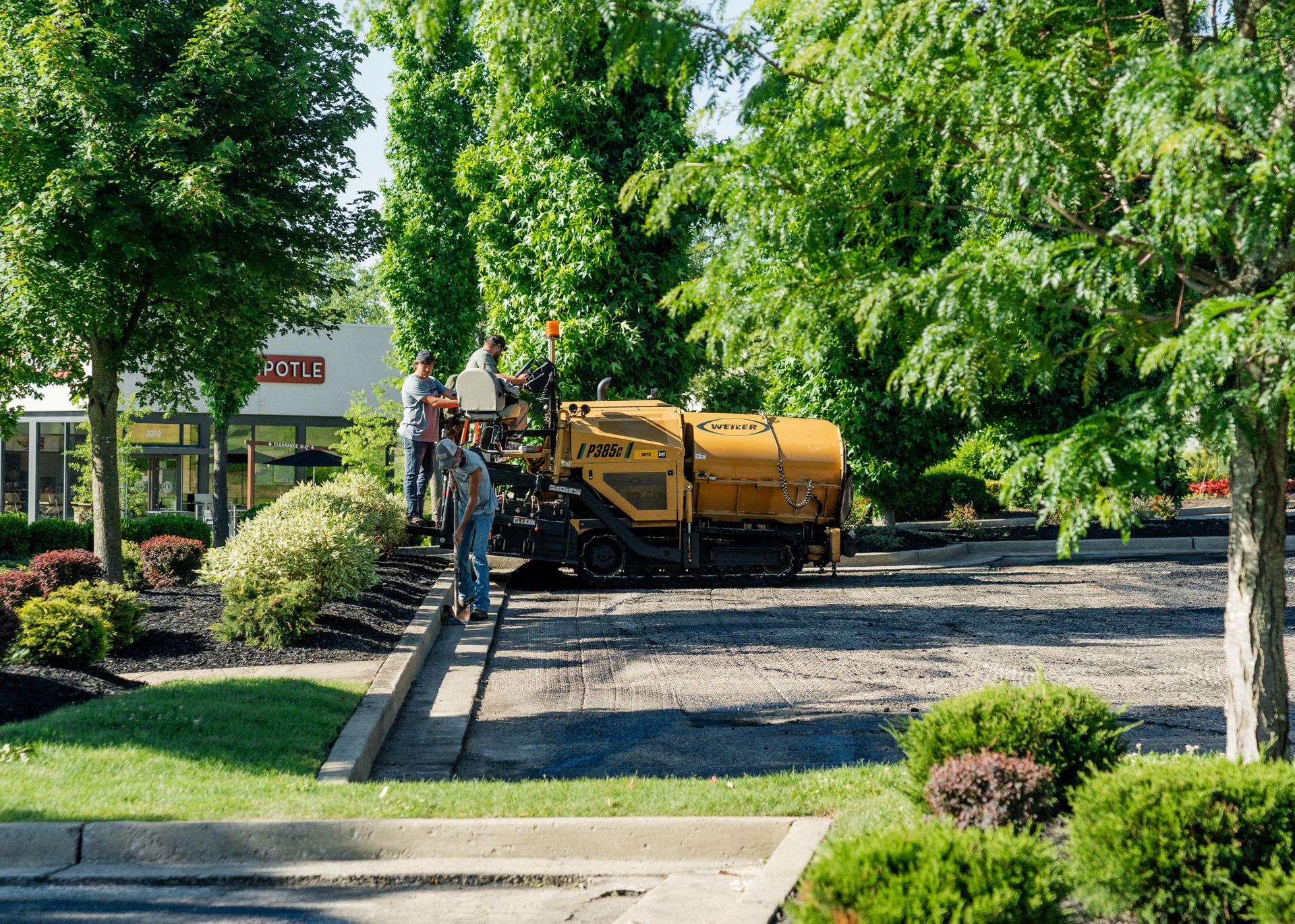 Yellow street sweeper on a leafy suburban road with workers nearby