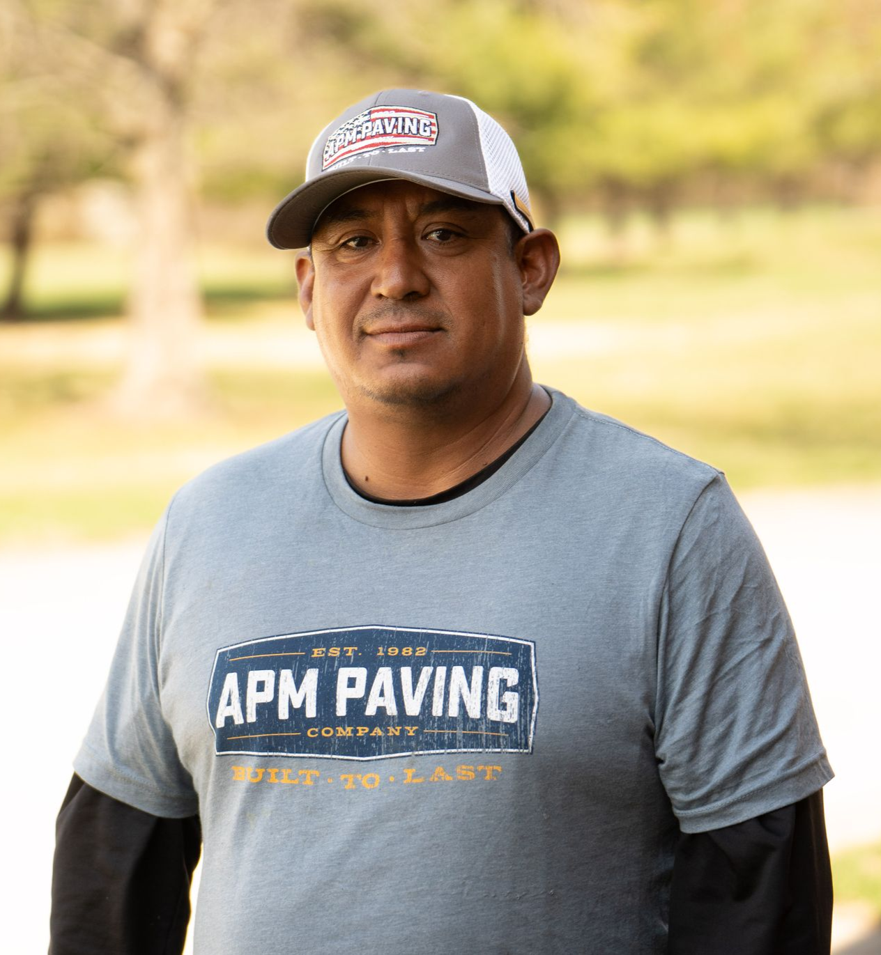 Man wearing an APM PAVING T-shirt and cap standing outdoors in a park-like setting