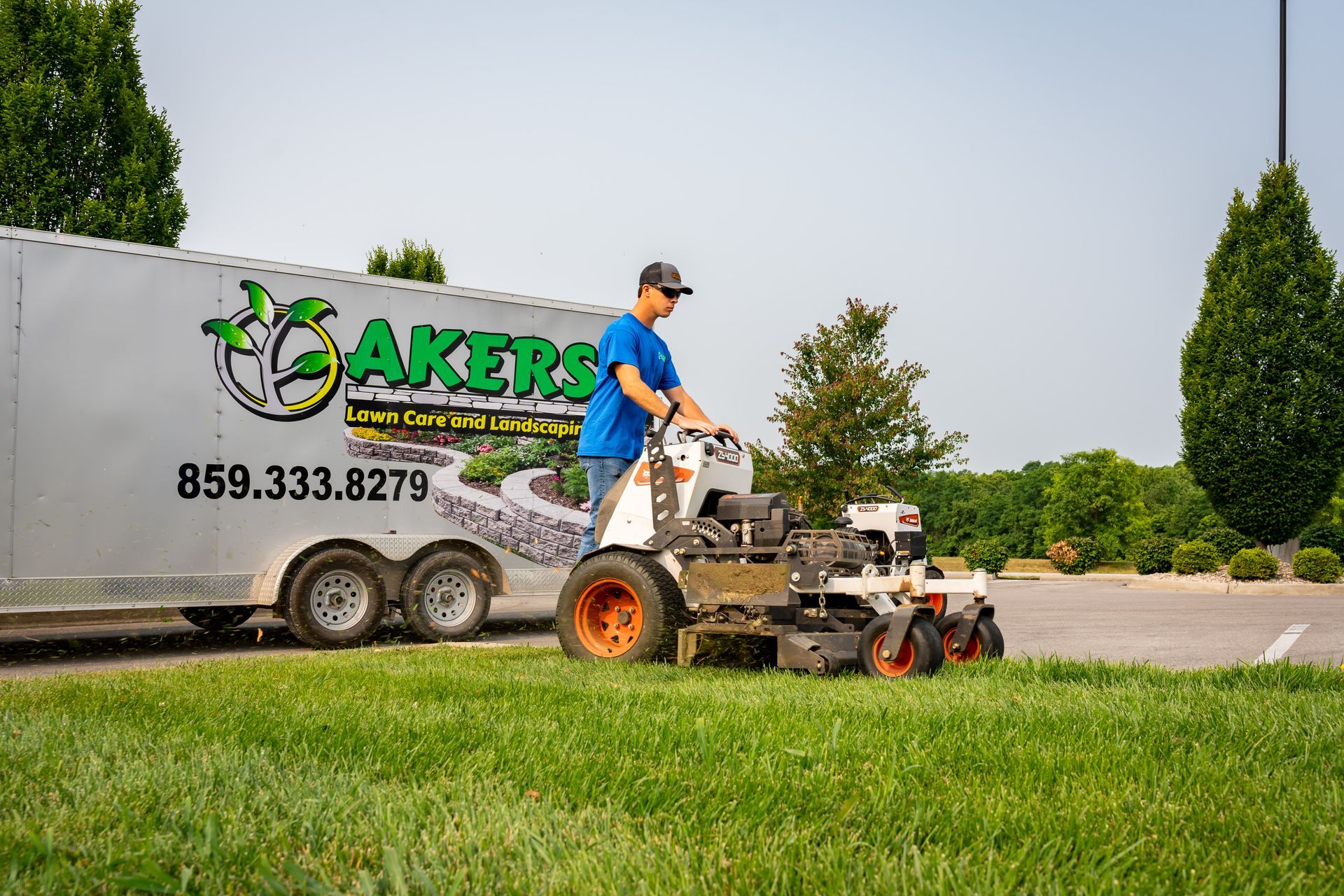 Landscaping worker mowing grass beside a company truck and trailer on a roadside lawn