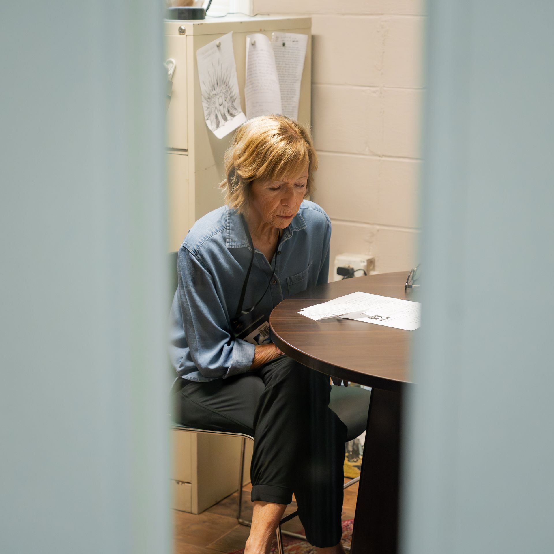Woman sitting at a table in a small room, looking down at papers with a thoughtful expression.