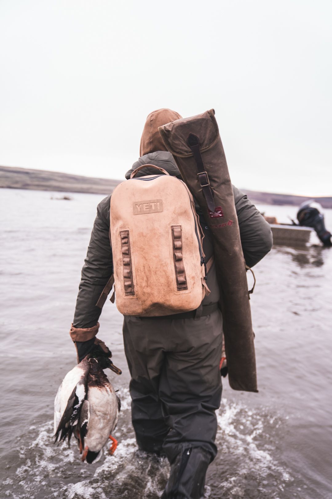 Person wades through shallow water carrying a backpack and a large fish, with a second person in the distance.