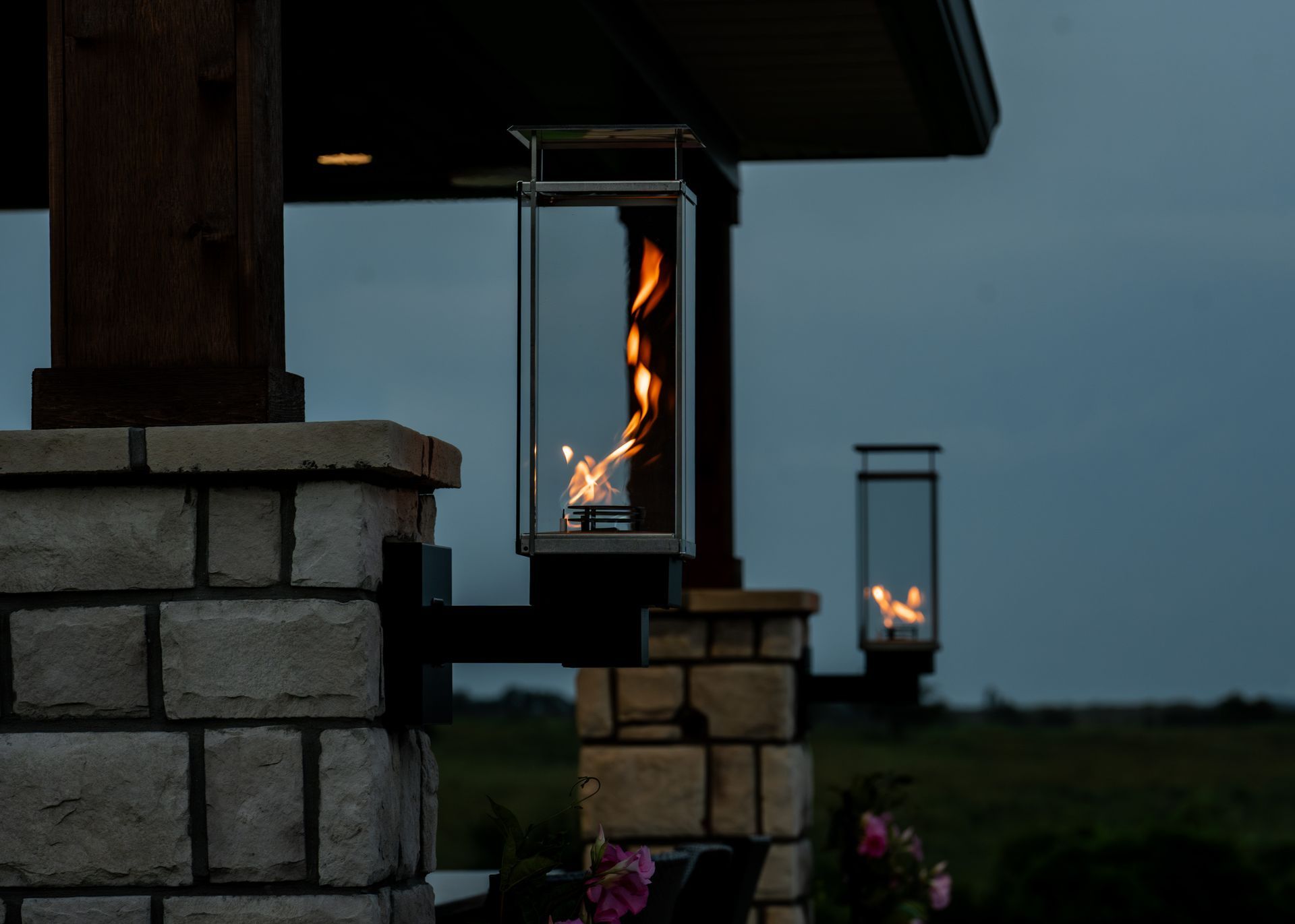 Lit lanterns hanging from a stone pillar at dusk, with one flame glowing in the foreground and another in the distance