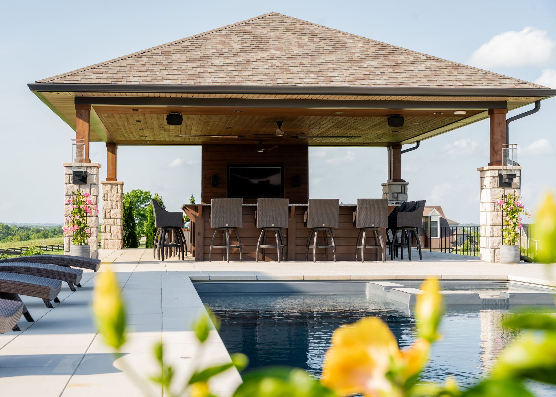 Poolside patio with a thatched-roof pavilion, dining table, and lounge chairs overlooking a bright landscape