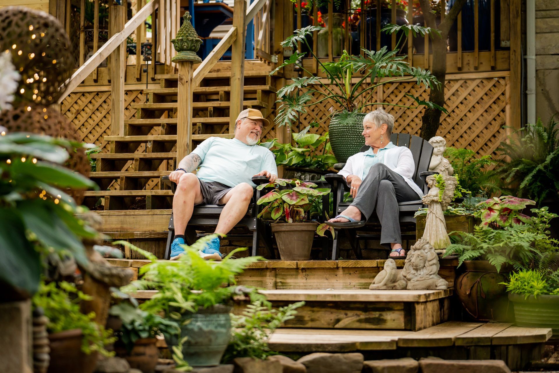 Two people sitting on chairs on a wooden deck in a lush garden, talking beside stairs and potted plants