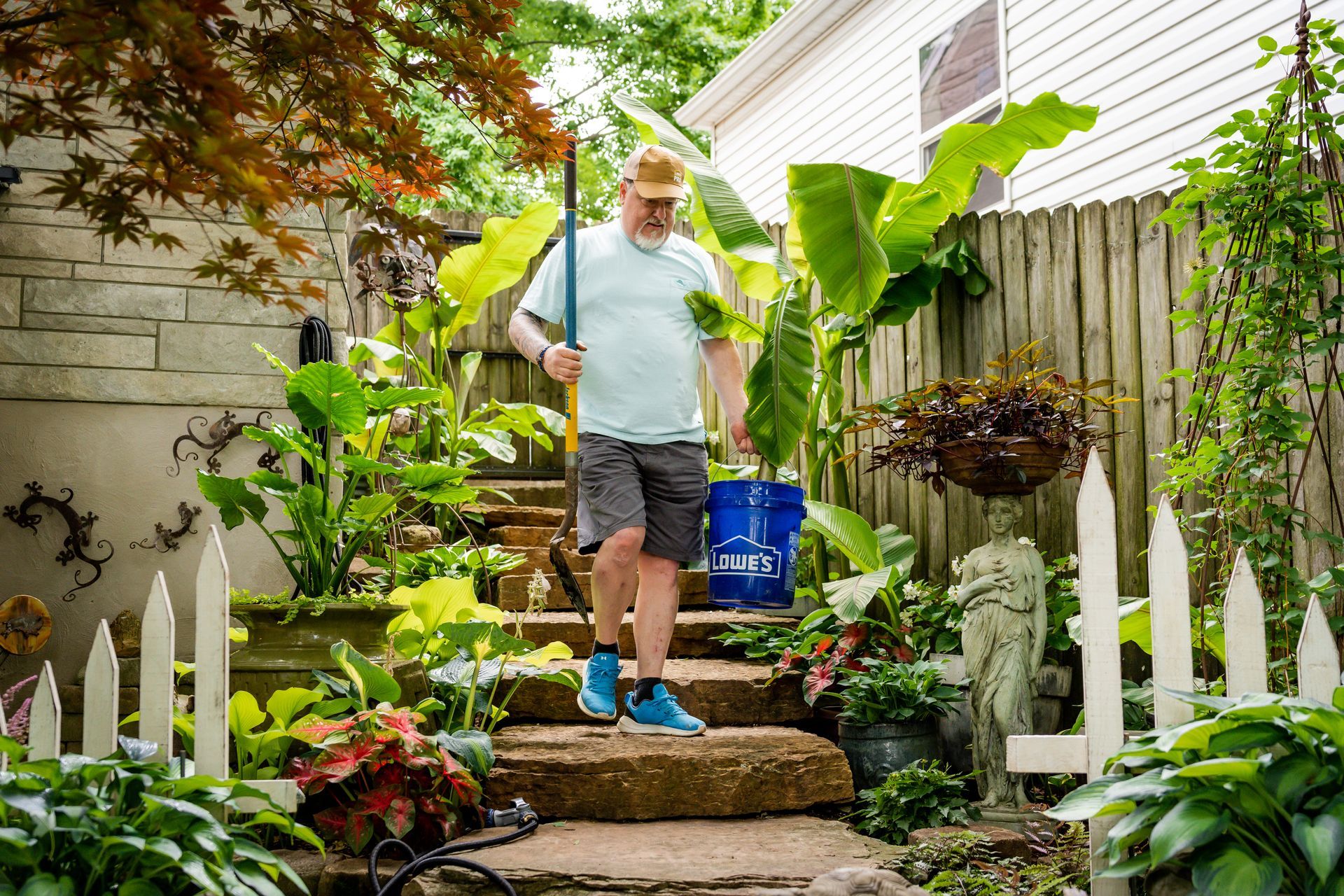 Gardener watering lush plants on stone steps in a backyard garden, carrying a blue bucket