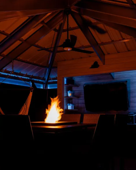 Cozy gazebo interior at night, lit by a glowing fire pit with dark chairs and a ceiling fan overhead