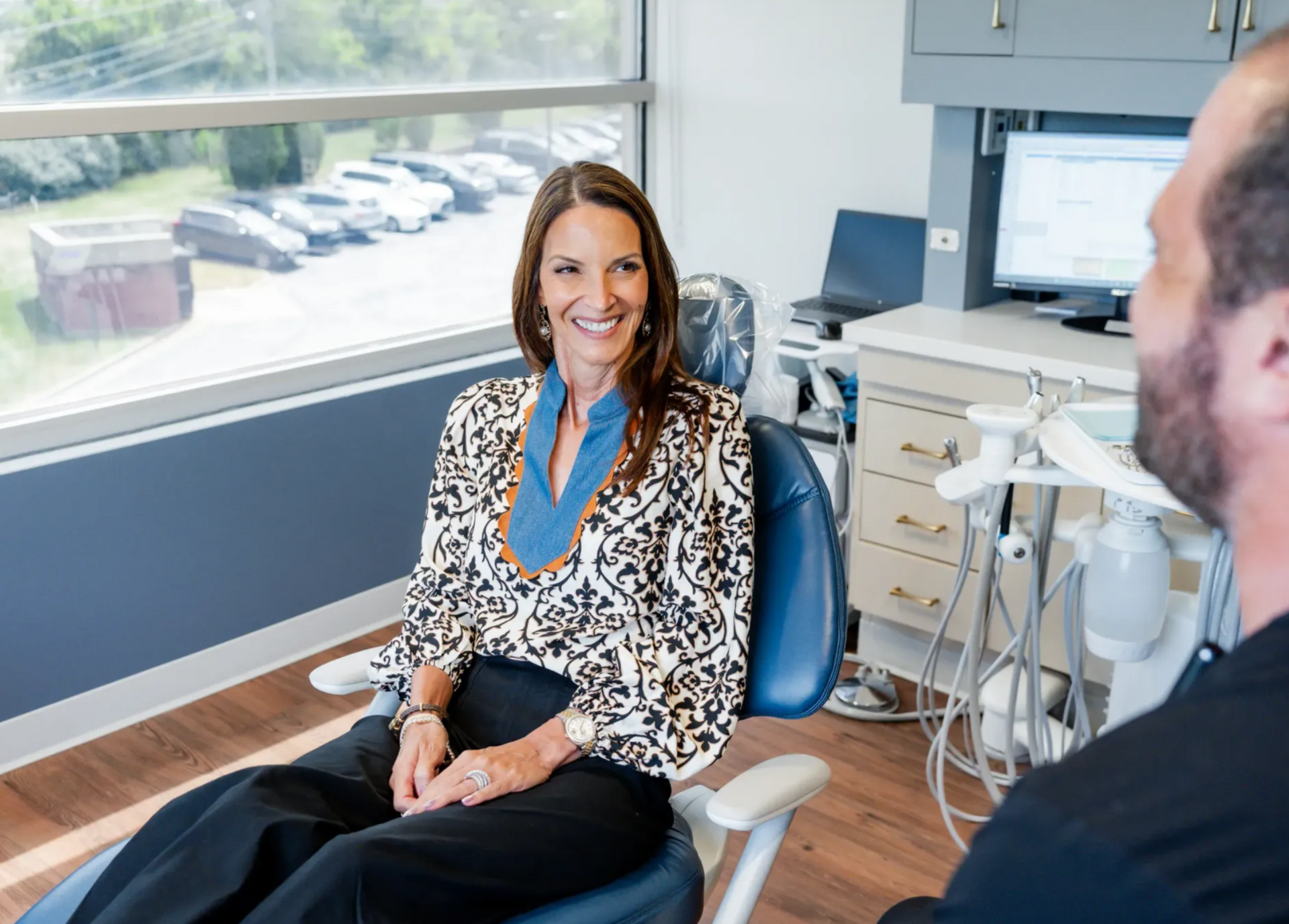 Smiling woman seated in a dental chair, talking with a man in a clinic office.