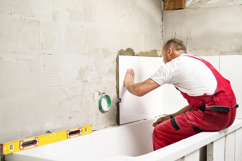 a Man is Sitting on a Bathtub While Installing Tiles on the Wall — Port Stephens Bathrooms In Nelson Bay, NSW