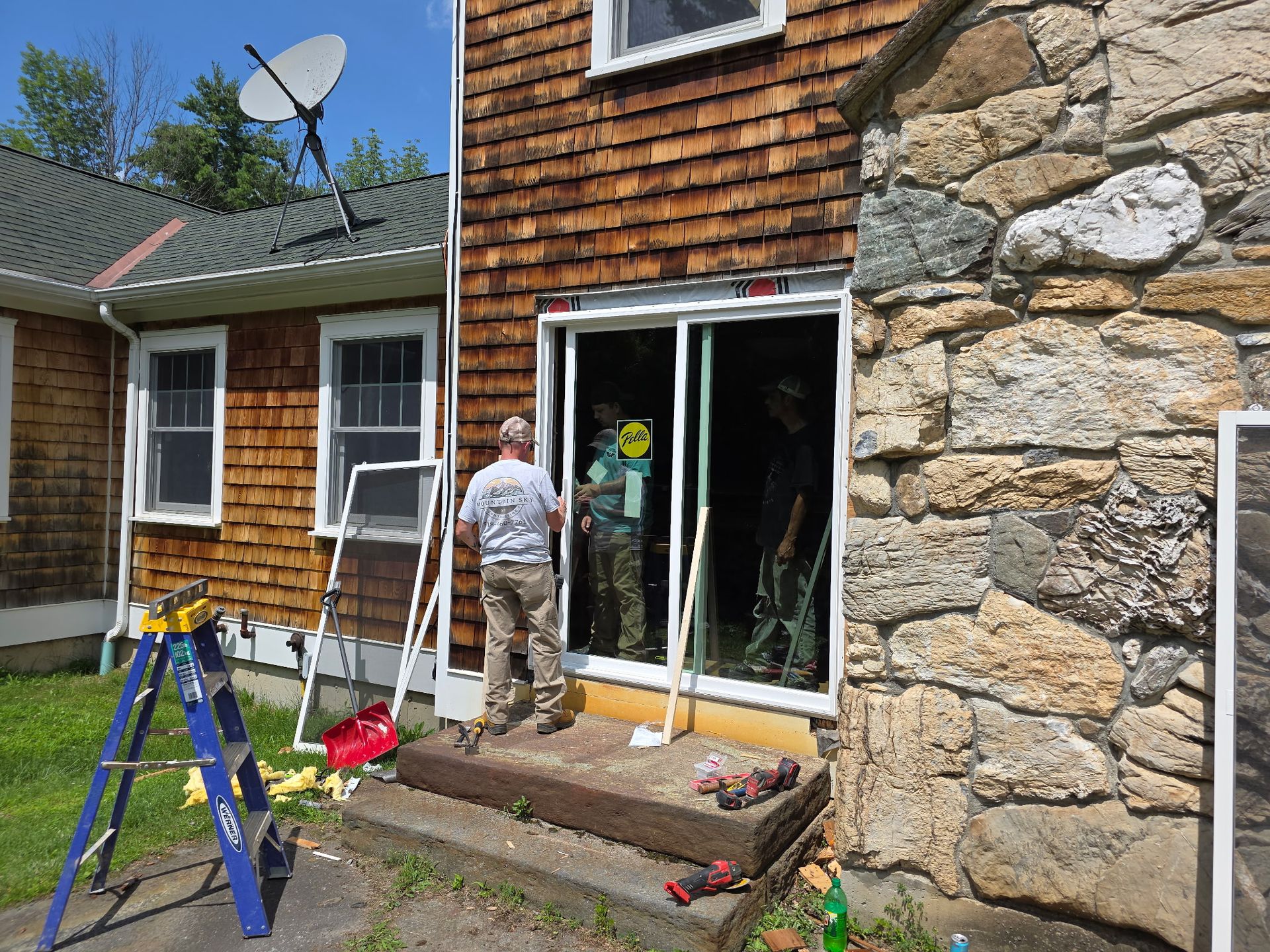 Workers installing a sliding glass door on a house exterior beside a stone wall and ladder