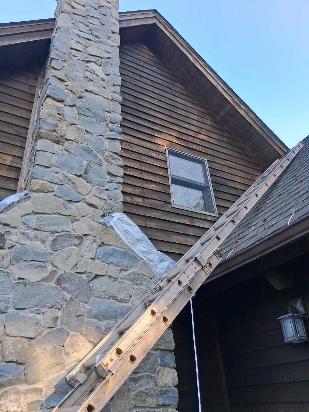 Wooden ladder leaning against a stone chimney on a cabin roof under a blue sky
