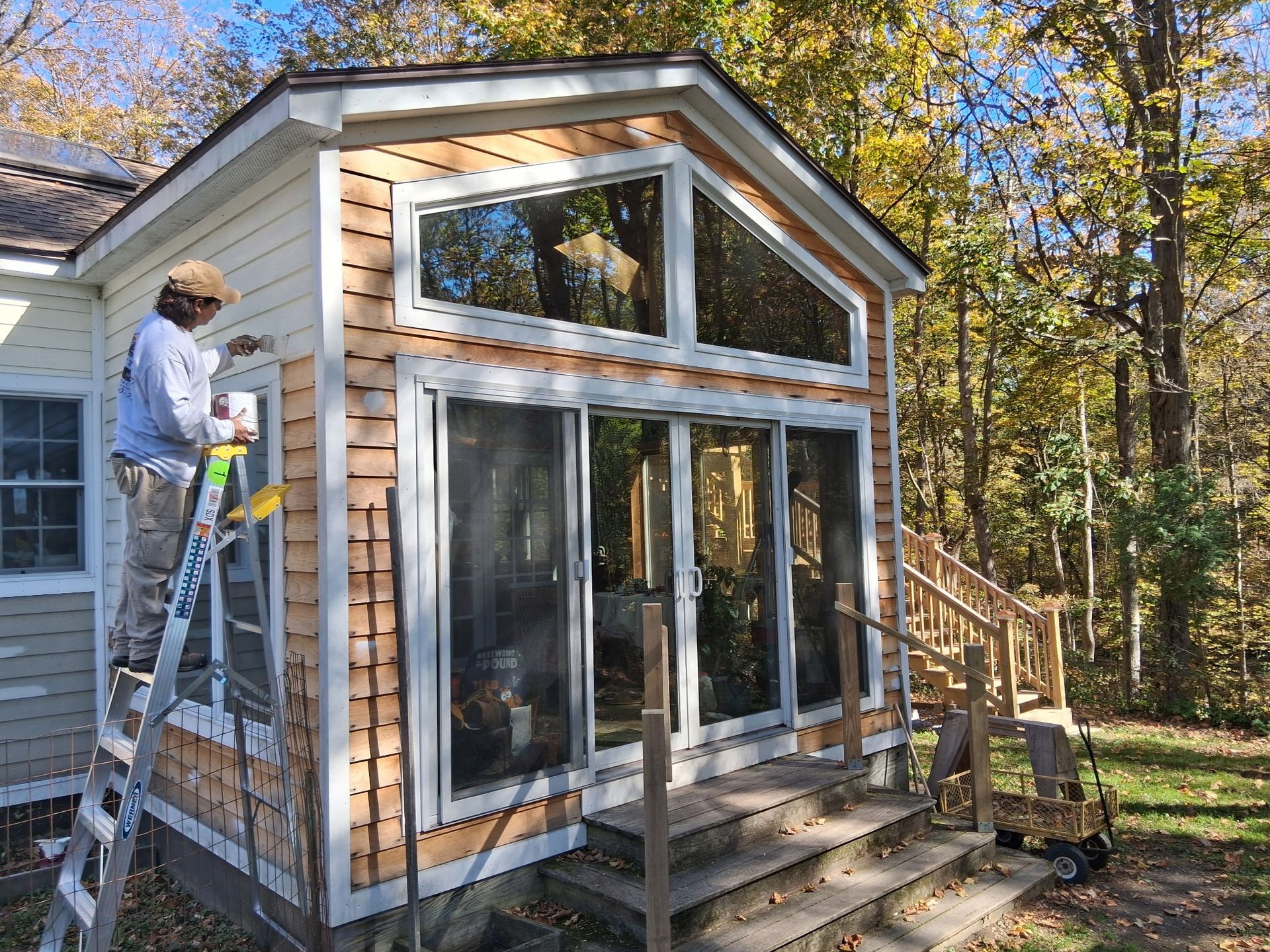 Worker on a ladder installing siding on a small house with large windows in a wooded yard
