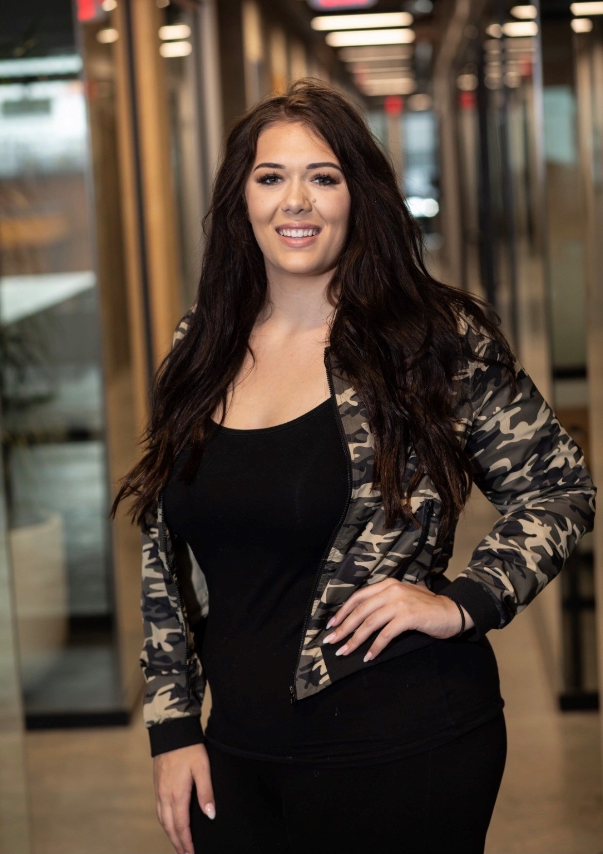 A woman in a black dress and a camouflage jacket is standing in a hallway.