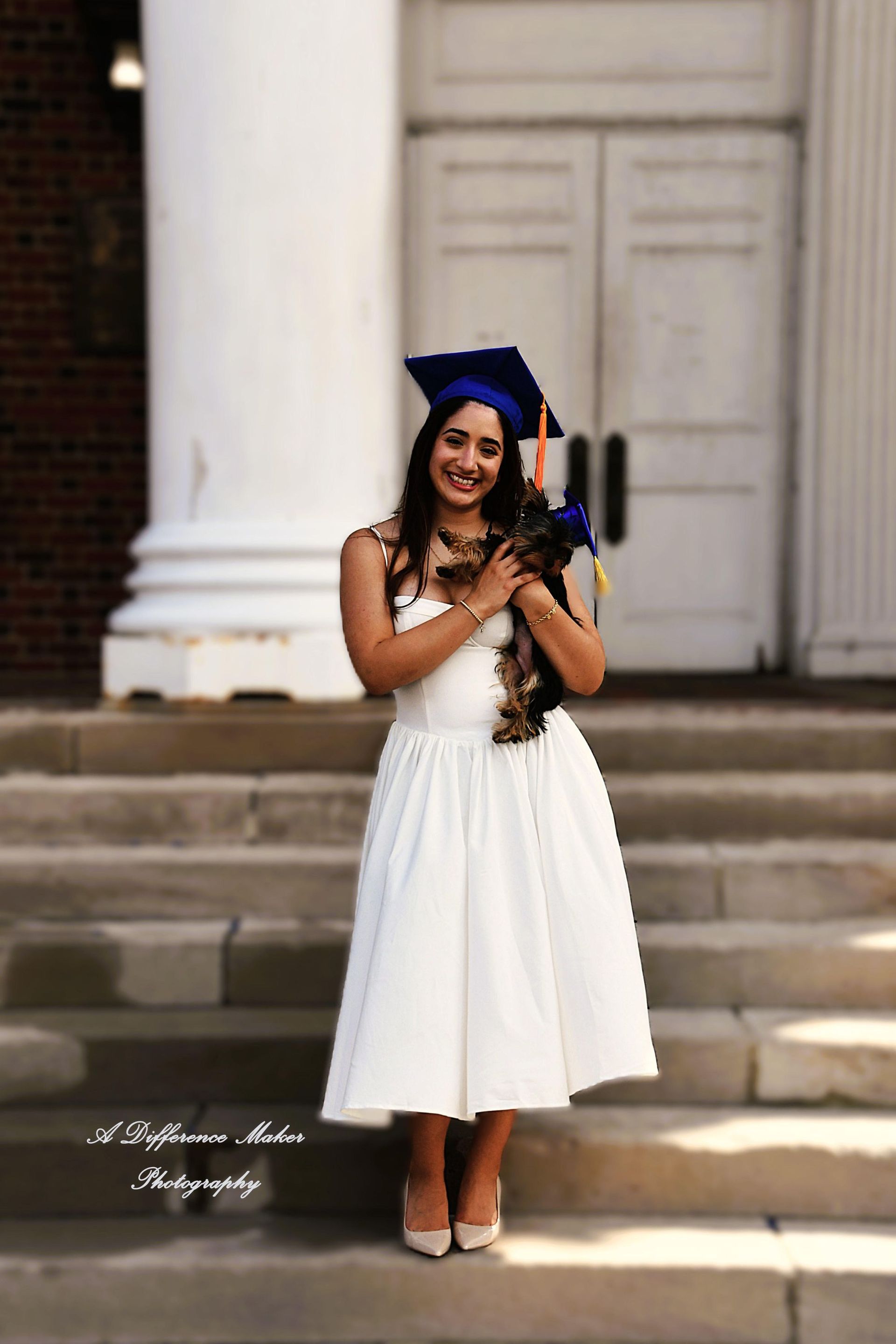 Woman in graduation cap and white dress holding a small dog, standing on steps in front of a building.