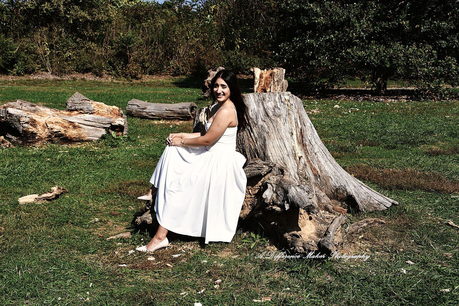 Woman in white dress and flower crown sits next to a tree stump on a grassy field.