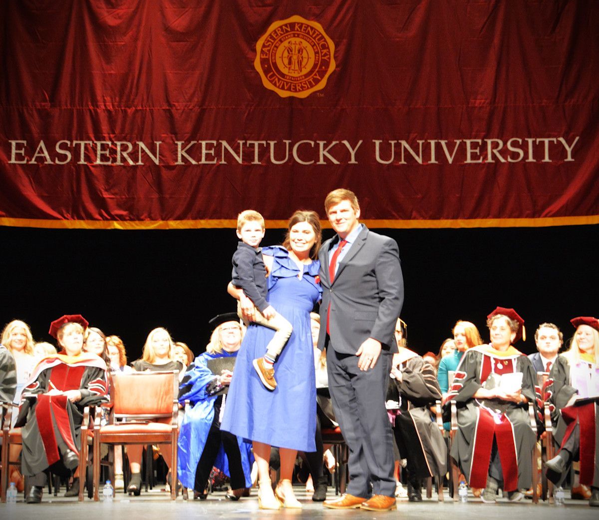 Family of three on stage at Eastern Kentucky University graduation. Woman in blue, man in suit, child held.