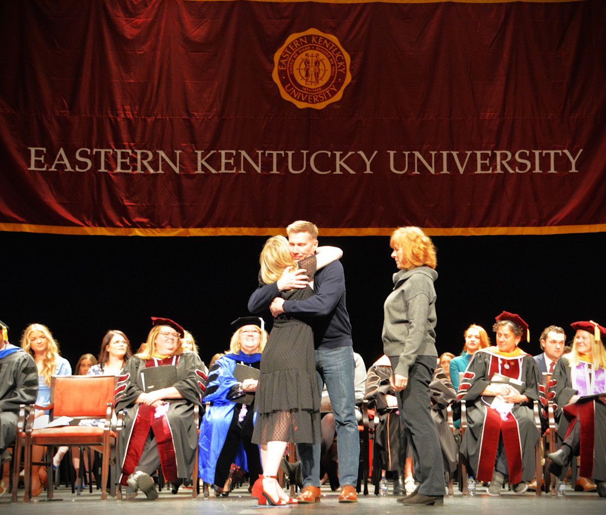 Eastern Kentucky University graduation: Man hugs a woman onstage. Another woman stands nearby.