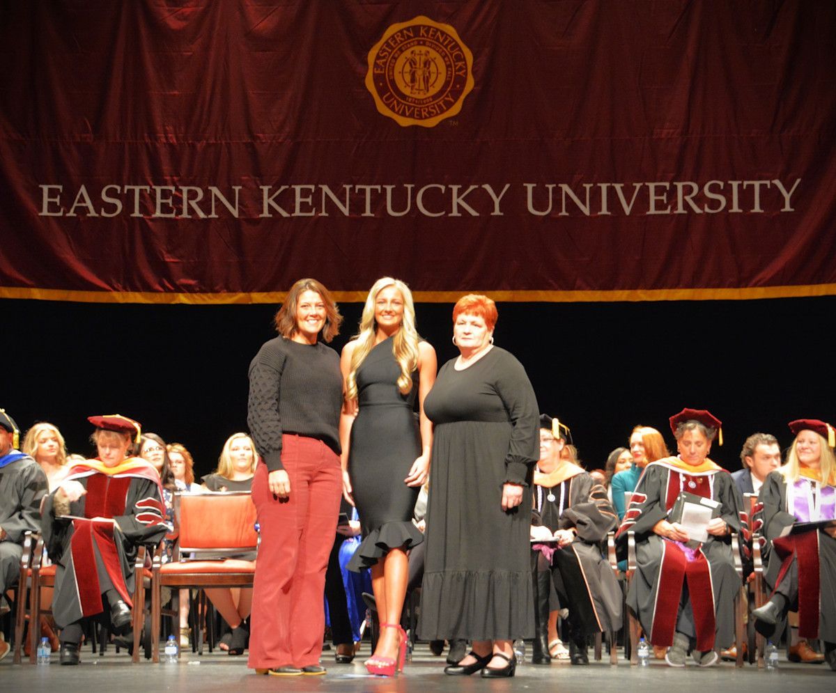 Three people on stage at Eastern Kentucky University graduation. They stand in front of a banner.