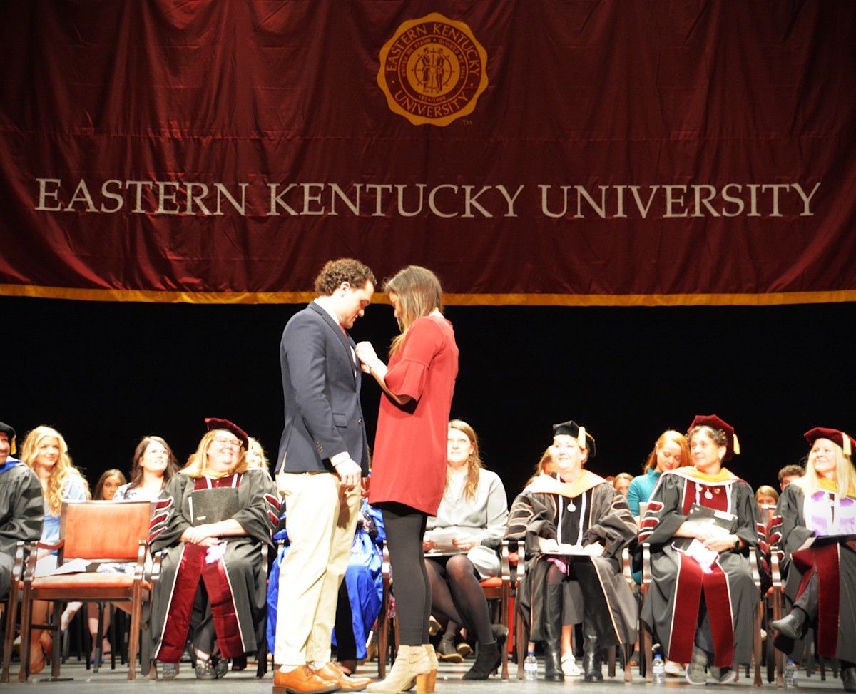 Man on stage being pinned by woman, in front of Eastern Kentucky University banner, faculty seated.