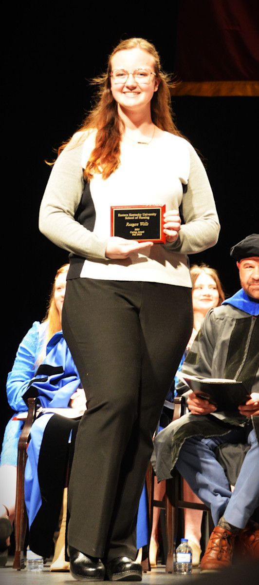 Woman in black pants and light-colored top holding an award on stage.