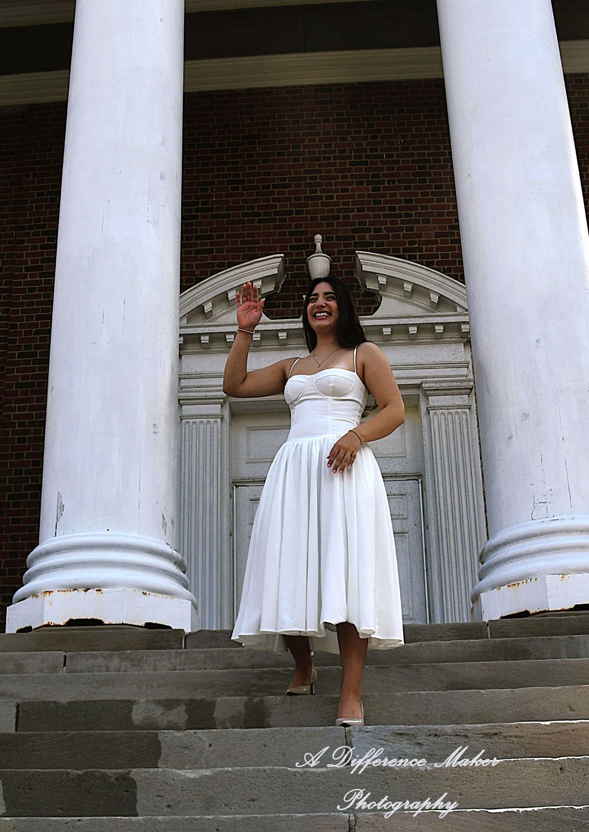 Woman in white dress waves from steps in front of a brick building with columns.