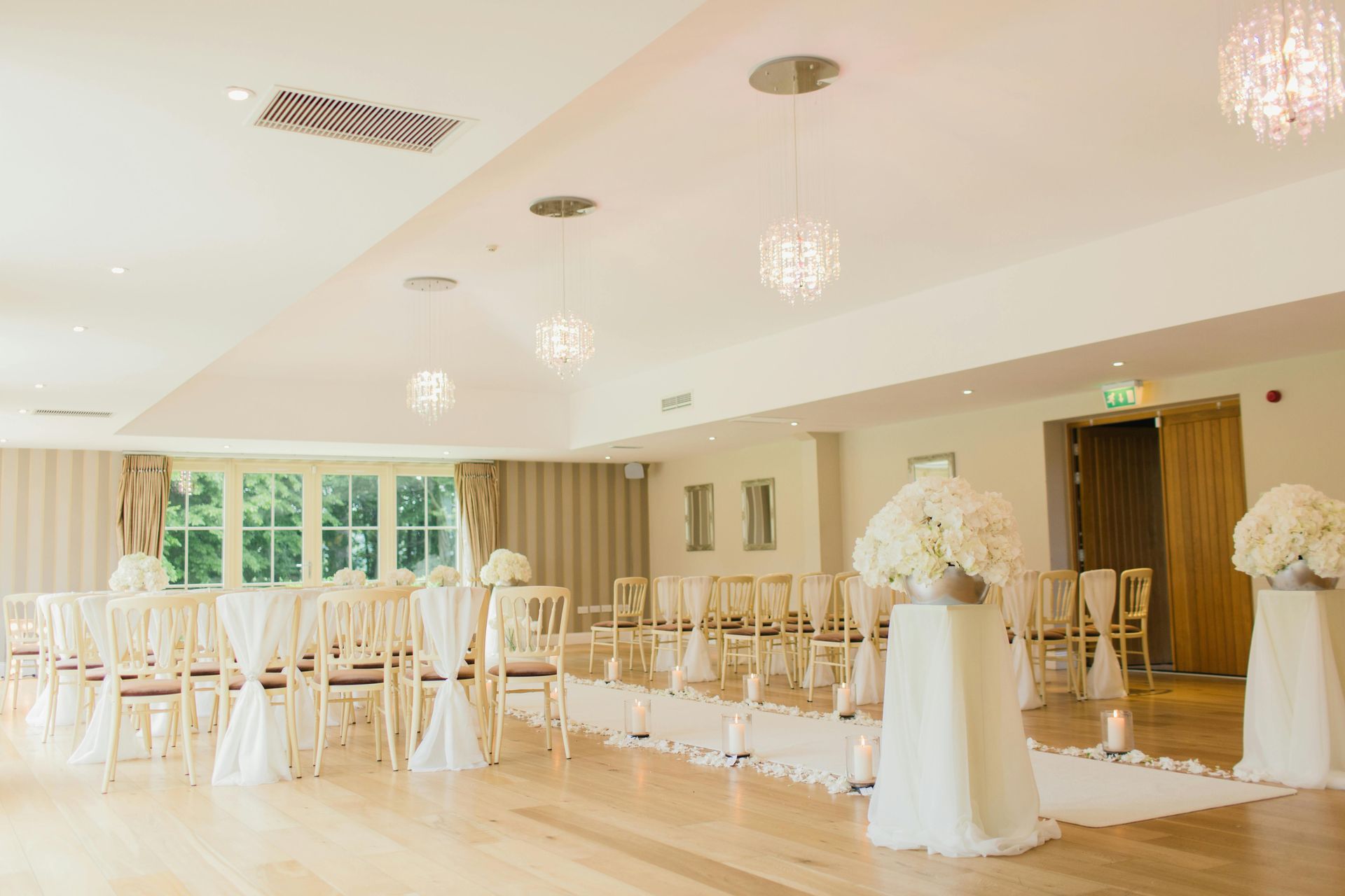 Wedding ceremony setup in a light-filled room with white floral arrangements, chairs, and decorative lighting.