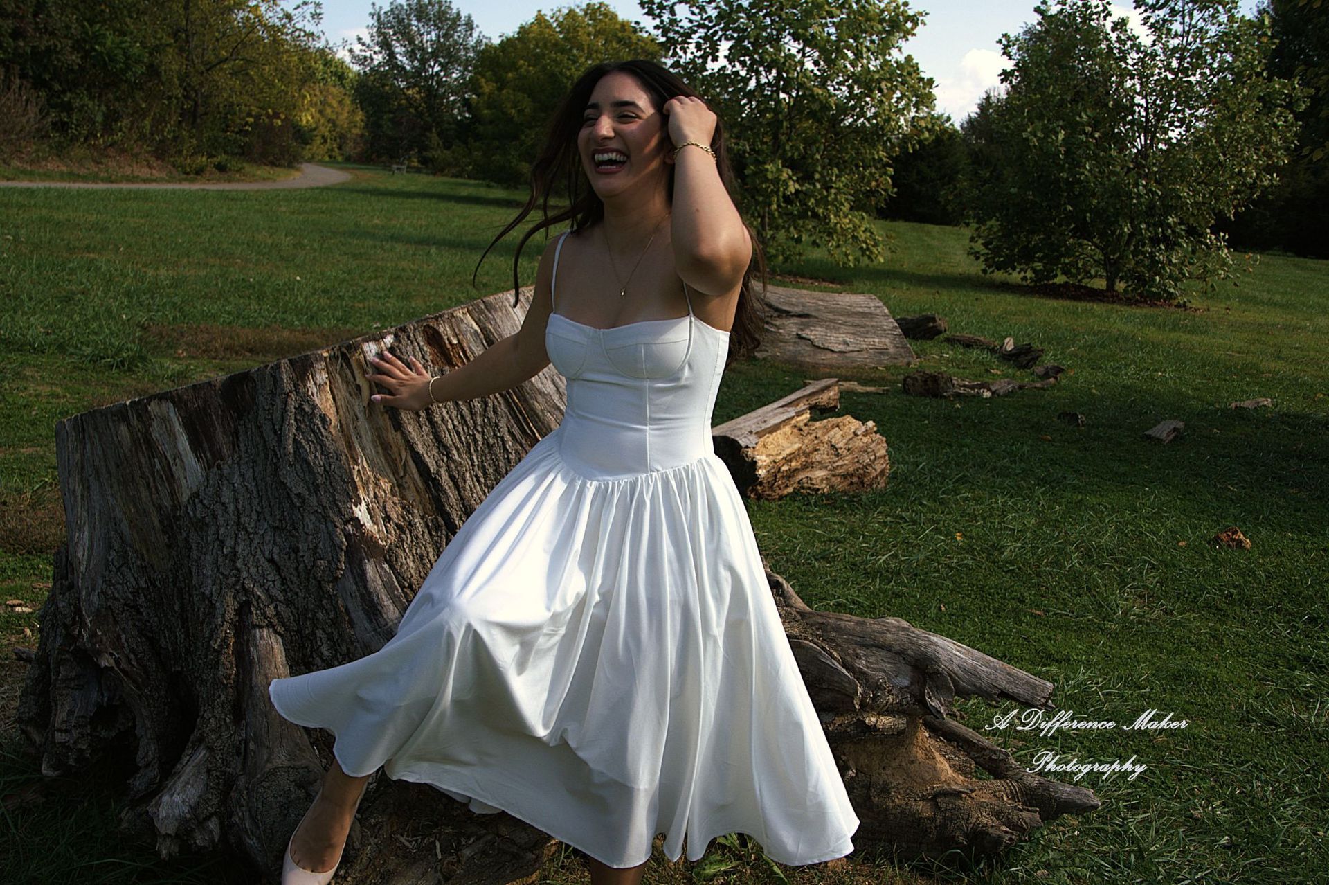 Woman in a white dress laughing outdoors, near a tree stump, with green grass and trees in the background.