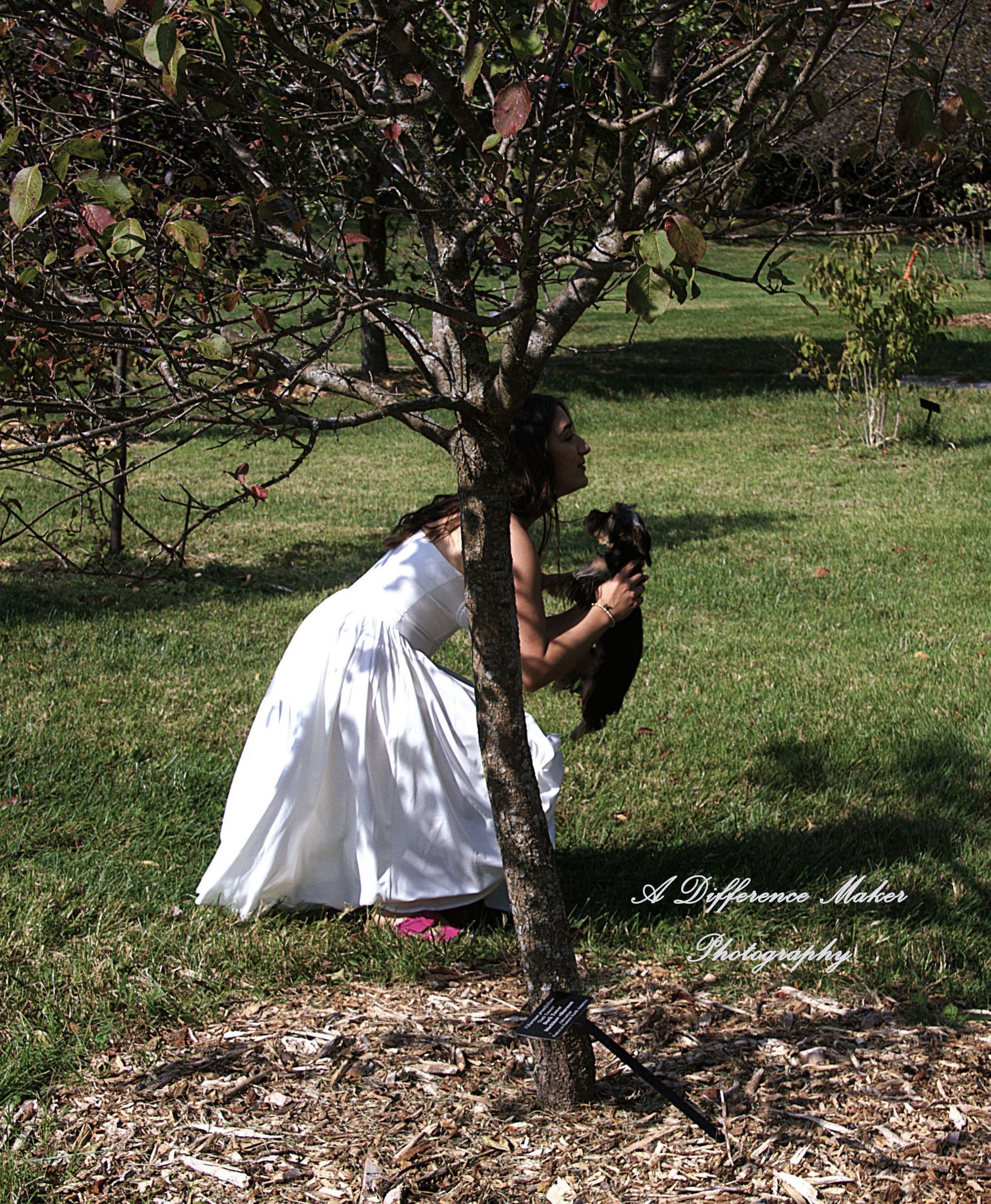 Woman in white dress and dog near a tree, outdoors. Dog is being held and kissed.