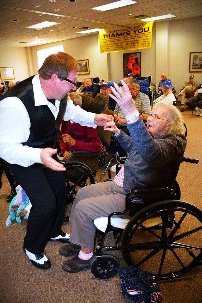 Man in vest dances with a person in a wheelchair at an event. Other people watch in a room with a