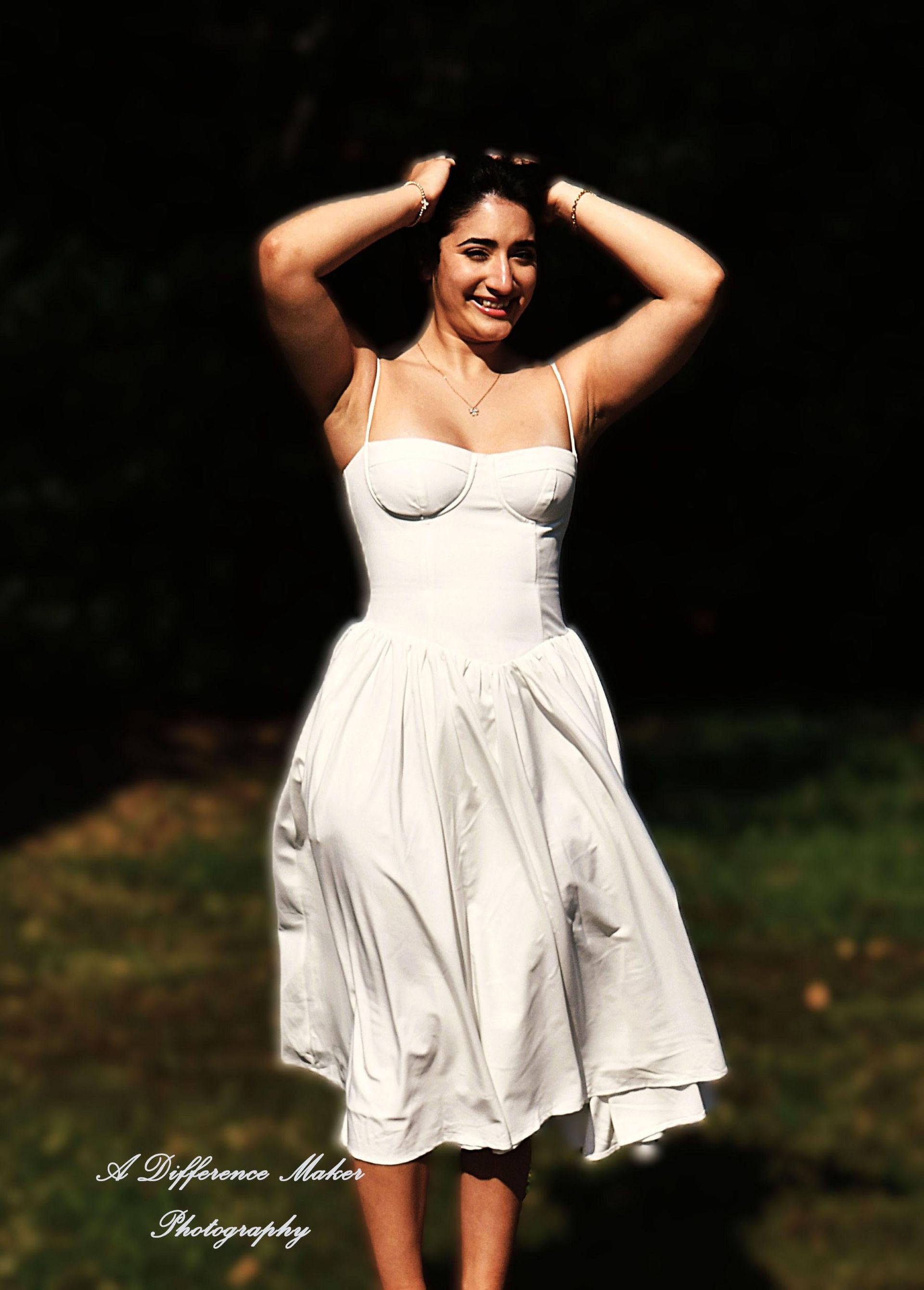 Woman in white dress with hands in hair, smiling in sunlight on grass.