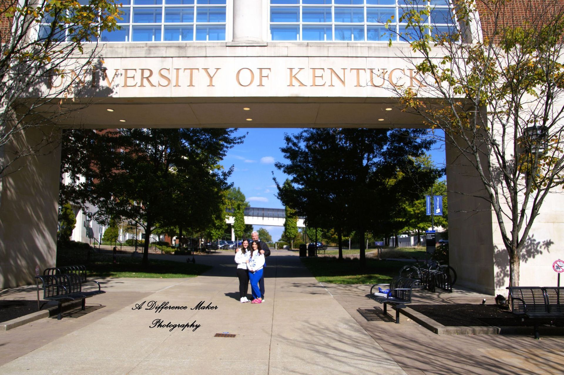 Entrance to the University of Kentucky, with two people standing under the archway. Blue sky and trees visible.